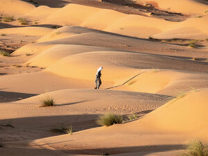 Against the backdrop of various sand dune formations, a traditionally dressed Omani wanders alone through the desert.