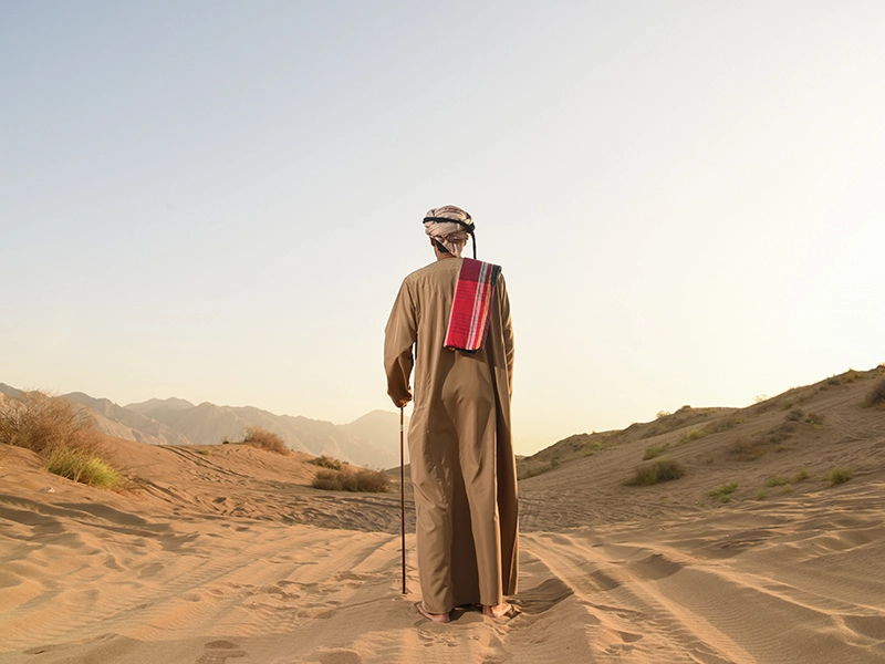 A man in traditional Omani attire walking through the sand dunes with mountains in the background