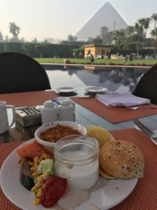 A table with plate of ful and yoghurt and view of pyramid in the background and water in the middle