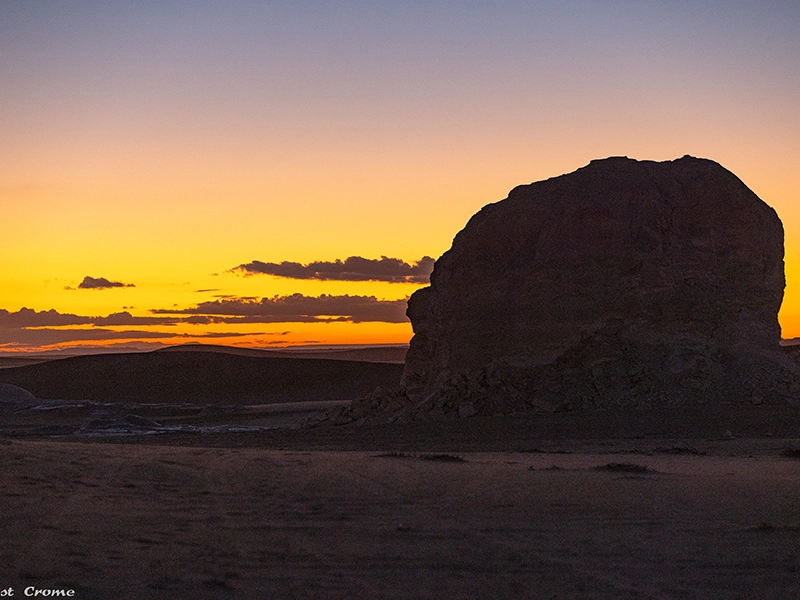 Der Sonnenuntergang in der Wüste färbt den Himmel rot und gelb, während ein Feld im Vordergrund nur als Silhouette zu erkennen ist.