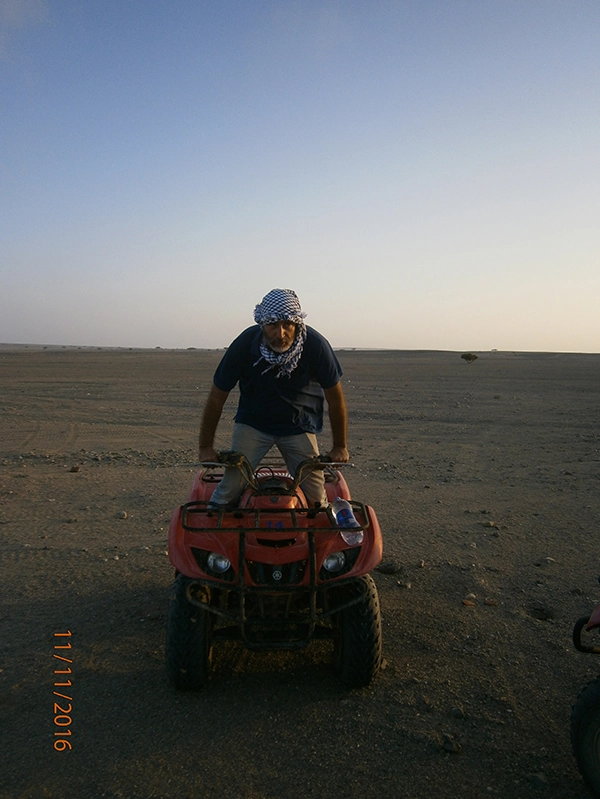 A man with white and black traditional headscarf in black t-shirt and khaki trousers standing on a stationary red quad bike in a field and clear blue sky