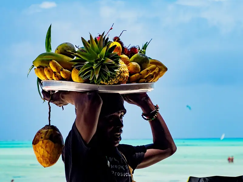 A local man walks along the beach in Zanzibar carrying a large plate of various fruits on his head.